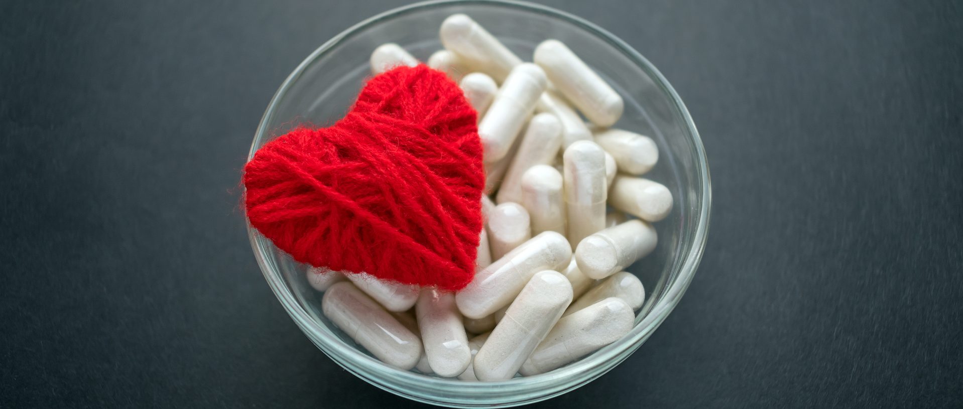 white veg capsules and red thread heart in glass bowl on black background, anticoagulant, blood thinners