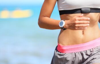 Runner woman with heart rate monitor running on beach with watch and sports bra top.
