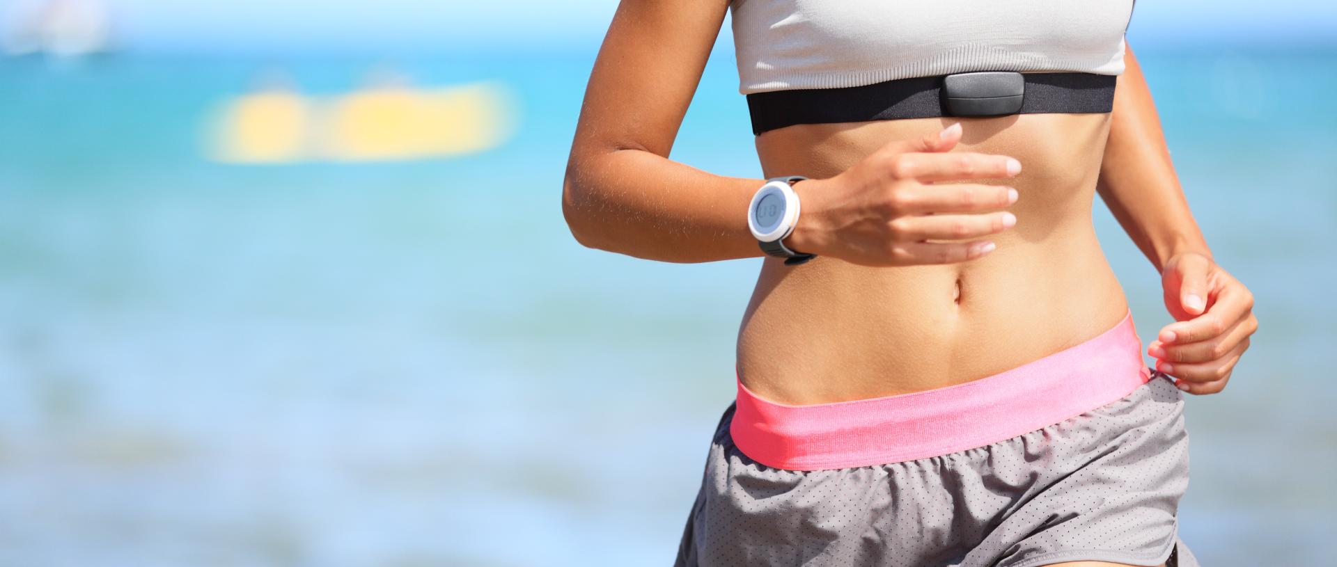 Runner woman with heart rate monitor running on beach with watch and sports bra top.