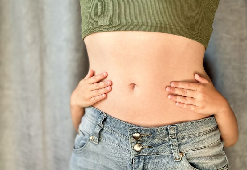 Un niño detrás de una mujer delgada con abdomen plano, abrazando a su madre. La mujer lleva jeans y un top corto verde.