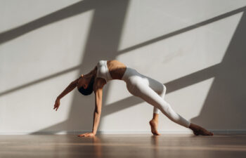 A woman in white athletic wear performs an advanced backbend, balancing on one hand and the ball of one foot on a polished wooden floor. Strong diagonal shadows are cast across the plain white wall behind her in a sunlit room.