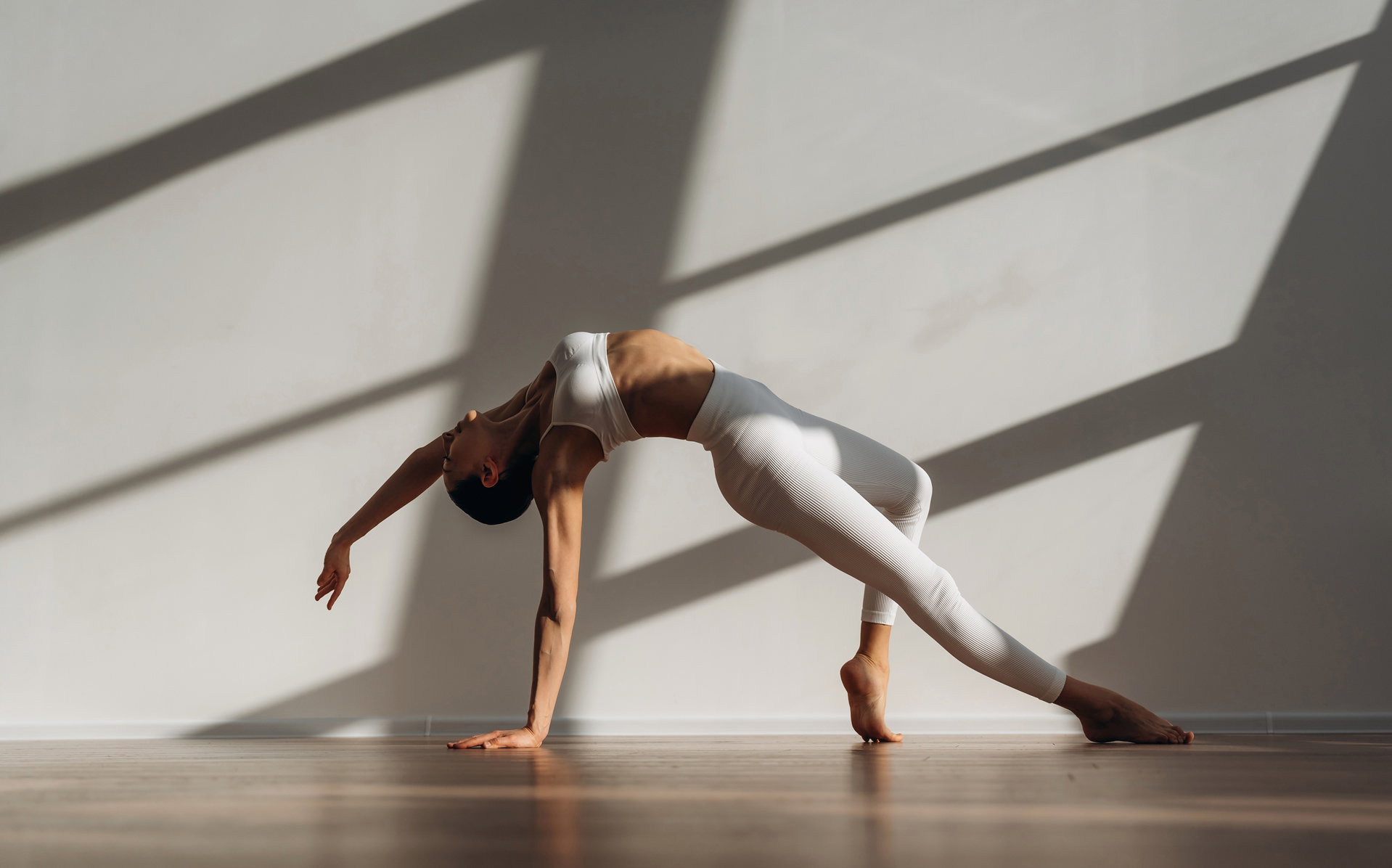A woman in white athletic wear performs an advanced backbend, balancing on one hand and the ball of one foot on a polished wooden floor. Strong diagonal shadows are cast across the plain white wall behind her in a sunlit room.