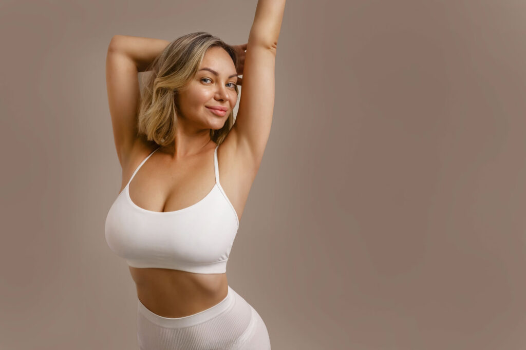 A confident woman with blonde hair smiling and posing with her arms raised behind her head. She is wearing a white sports bra and matching high-waisted leggings against a solid beige background.