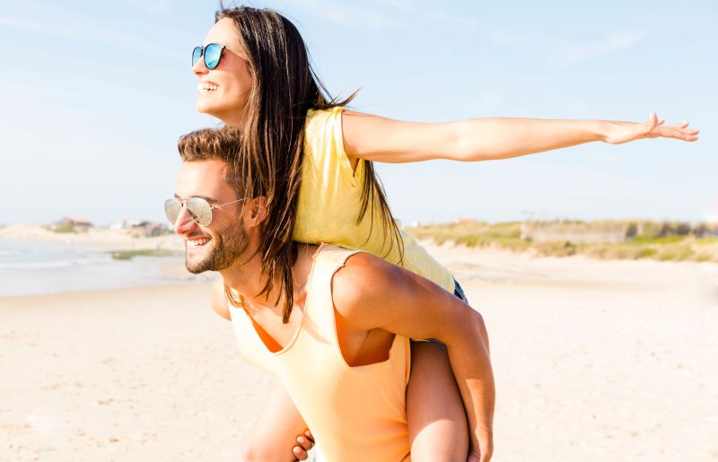 A happy couple spending time on a beach. Athletic handsome man piggybacking a smiling slim woman who 