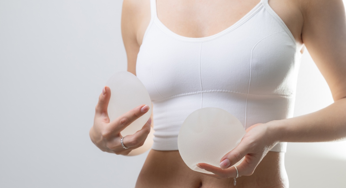 Person wearing a white sports bra holds two silicone breast implants, one in each hand, against a neutral background.